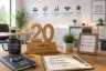 Schreibtisch mit einem hölzernen Jubiläums-Aufsteller '20 Jahre', daneben eine Tasse mit der Aufschrift 'Ich mache einen Unterschied', ein Notizbuch mit 'Weiterhin. Mit Freude. Mit Wirkung.' und Icons für Vertrauen, Wirkung, Entwicklung und Verantwortung an der Wand.