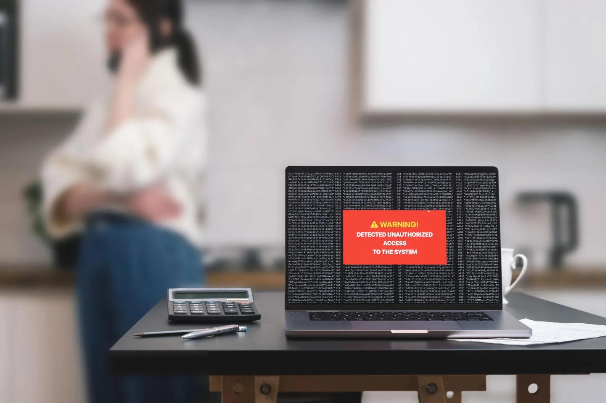 Laptop auf einem Küchentisch mit roter Warnmeldung 'Detected unauthorized access to the system', im Hintergrund unscharf eine telefonierende Person.