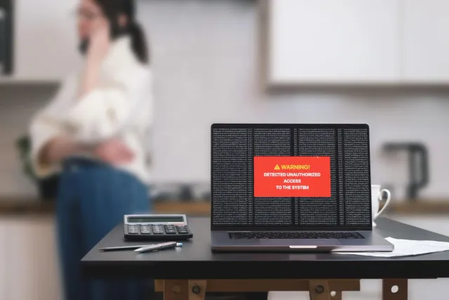 Laptop auf einem Küchentisch mit roter Warnmeldung 'Detected unauthorized access to the system', im Hintergrund unscharf eine telefonierende Person.